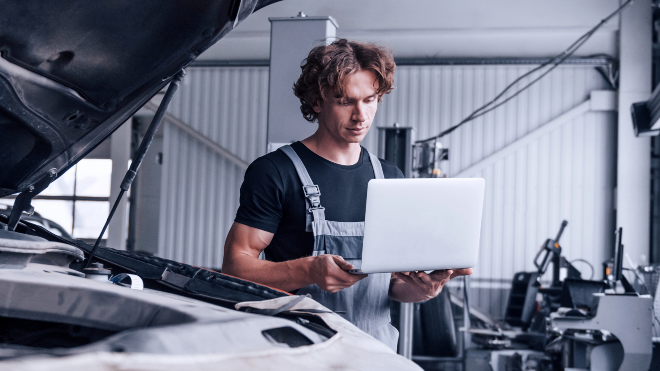 Mechanic holding a laptop beside a car with an open bonnet.