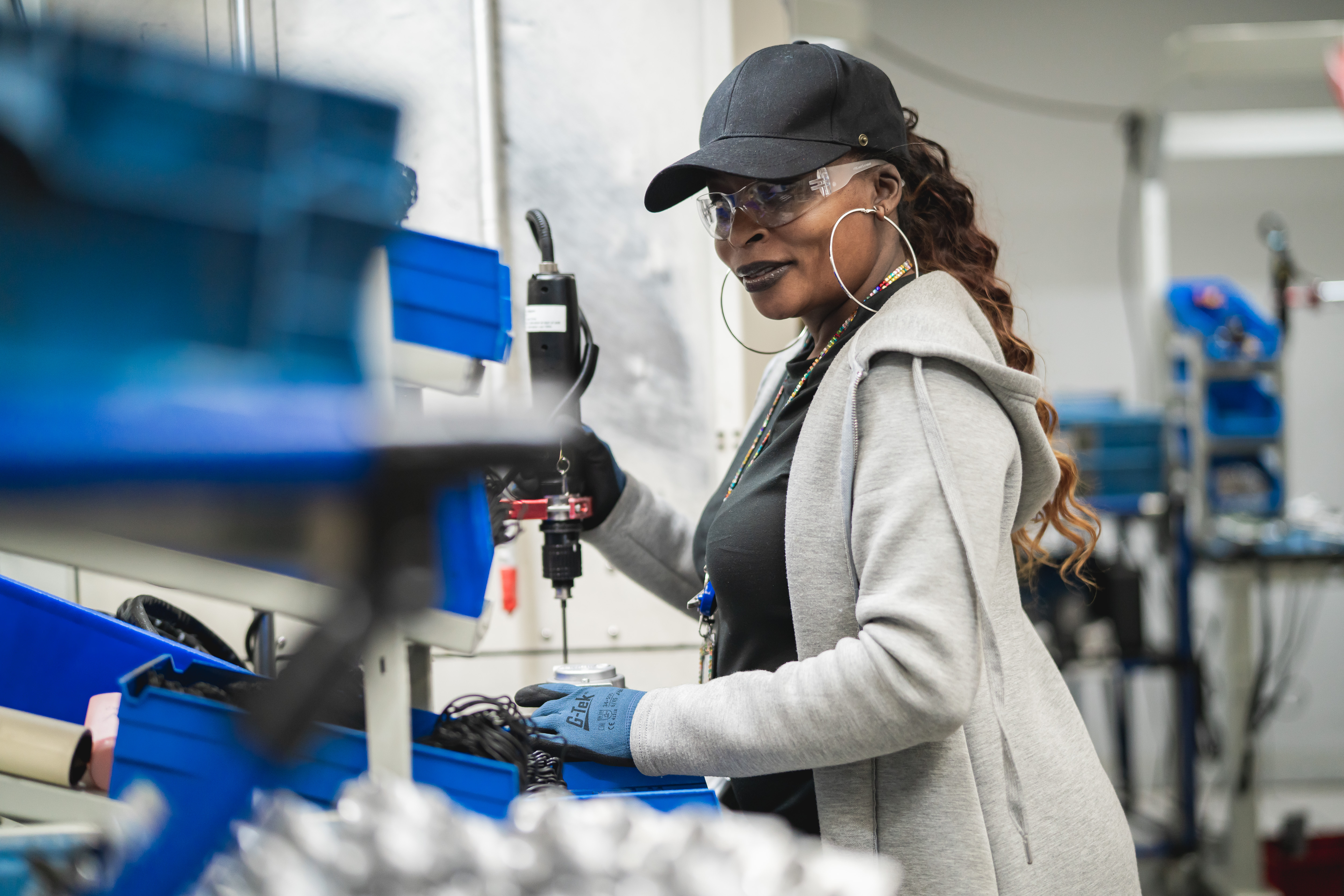 Female factory worker operating machinery while wearing safety goggles, gloves, and a black cap in an industrial setting