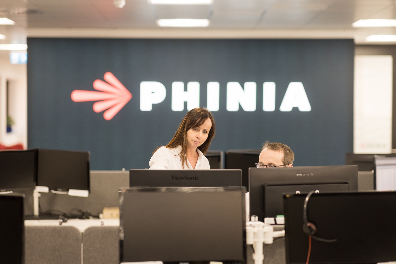 PHINIA employees collaborating in a modern open office workspace with a prominent PHINIA brand logo in the background.