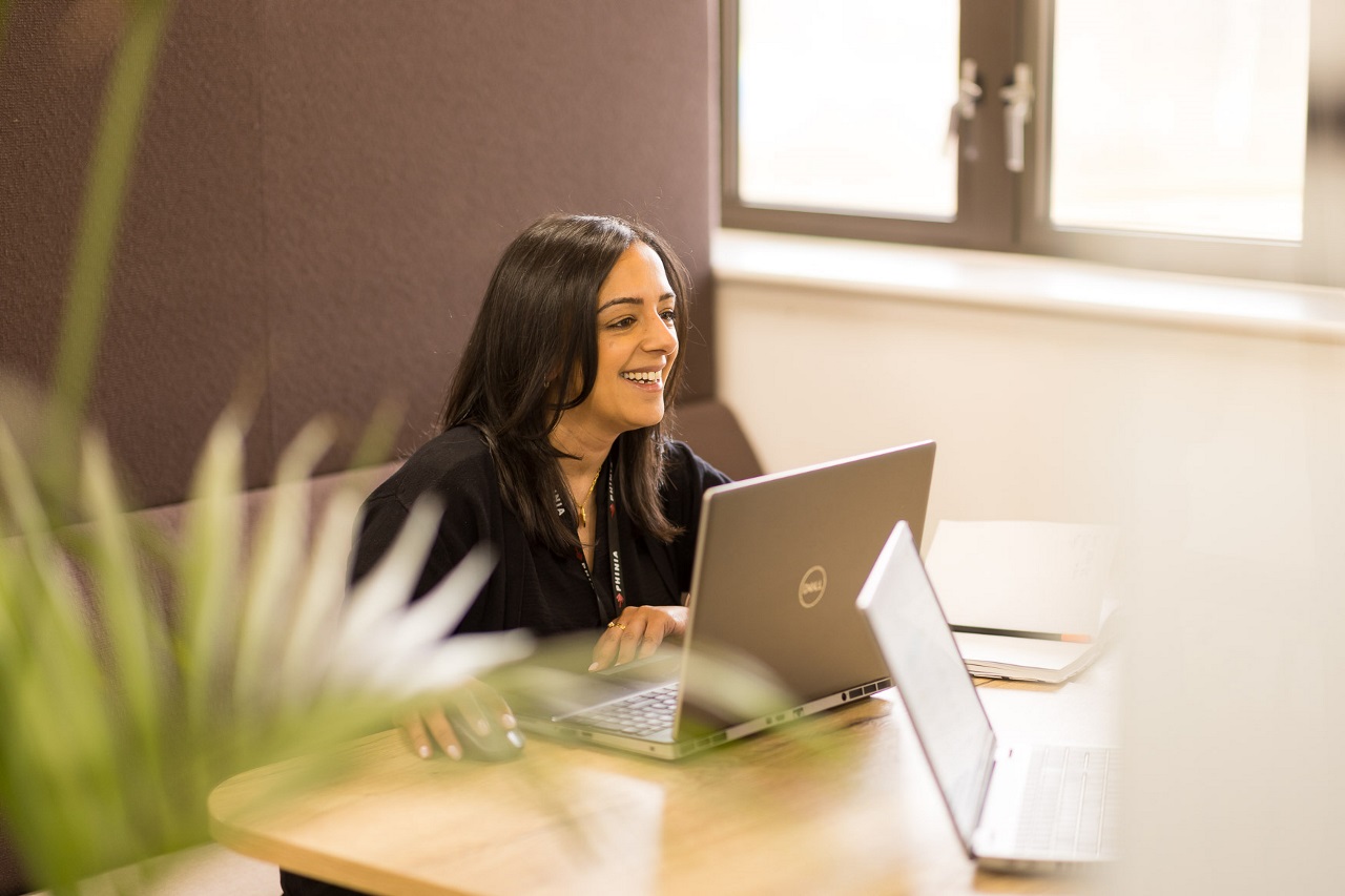 Woman smiling while working on her laptop at a small table in an office space, surrounded by natural daylight.
