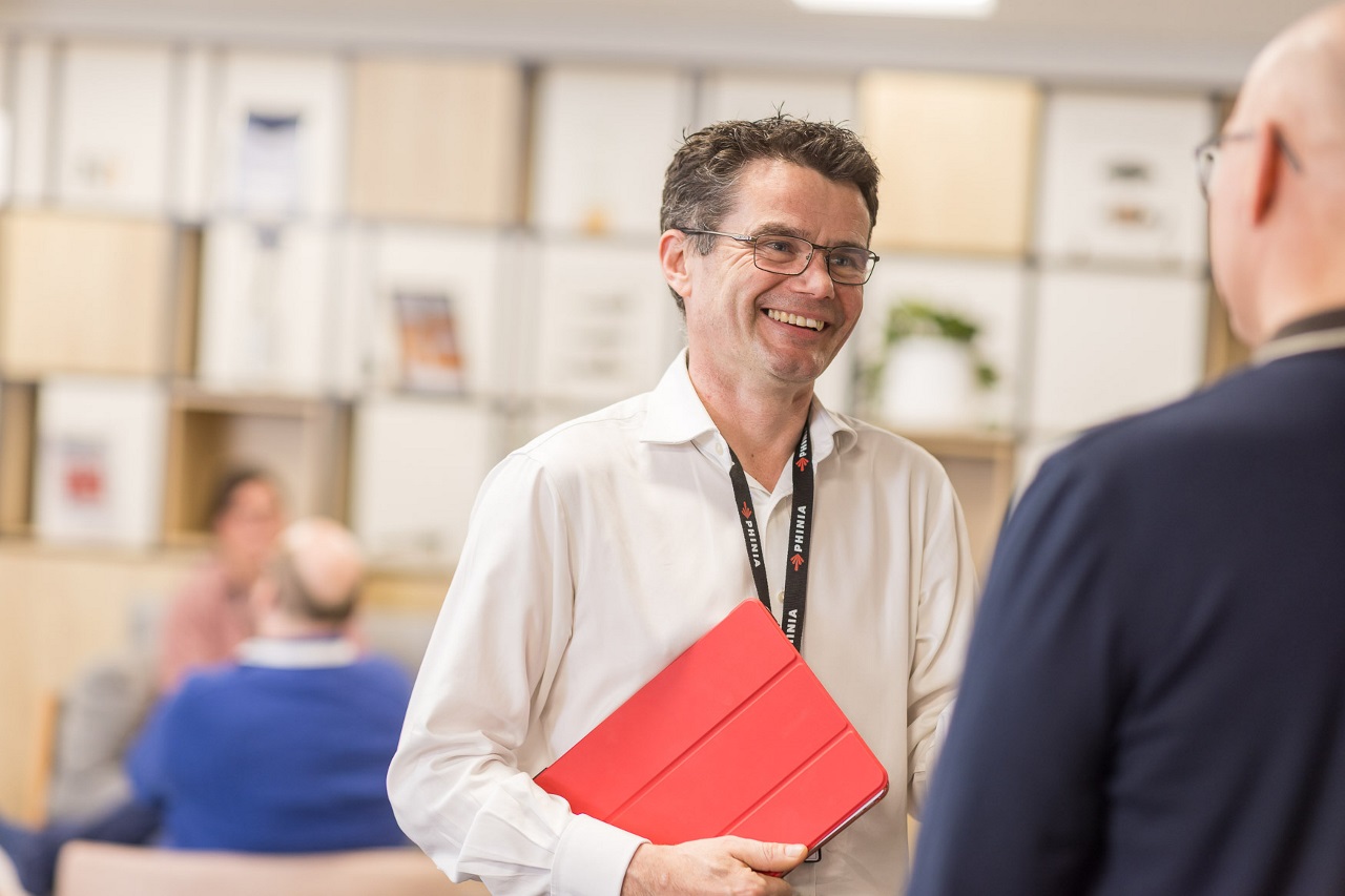 PHINIA executive smiling during a casual office discussion while holding a red tablet, reflecting openness and leadership approachability.